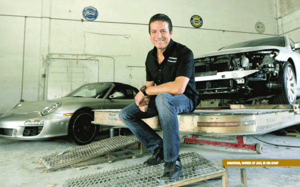 Jonathan Augusto sitting on a flatbed in the shop with a silver Porsche behind him and a wrecked white Porsche on the right